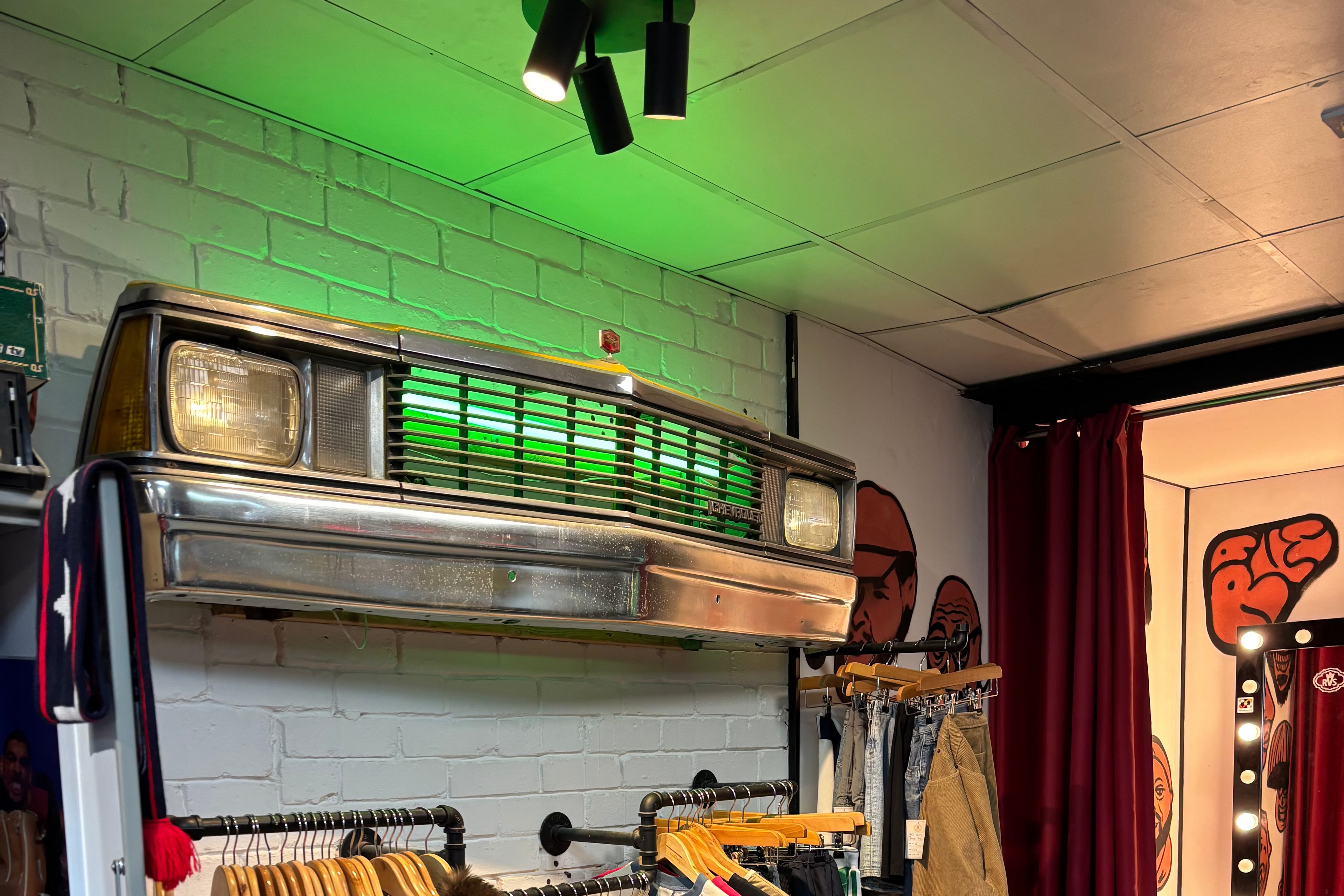 A clothing boutique interior with racks of garments hanging on wooden hangers, a brick-patterned wall, a vintage car grille decor piece, red curtains, and a leather chair in the foreground.
